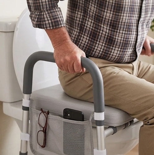 A close-up of a person sitting on a gray toilet training seat with armrests, holding the handles. The seat has a mesh side pocket holding eyeglasses and a smartphone, with the person wearing a brown plaid shirt and khaki pants.