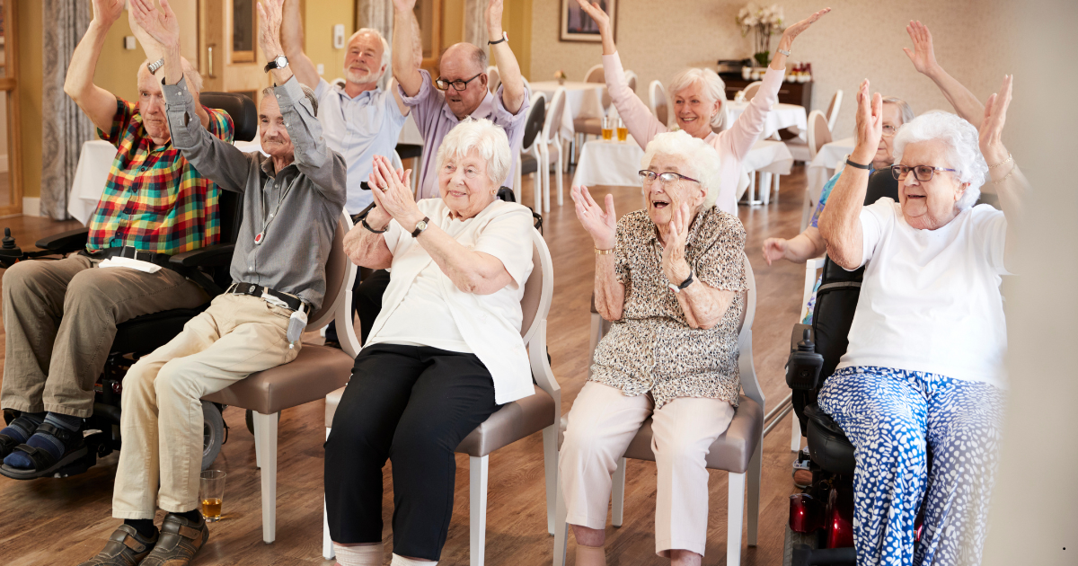 A group of elderly people sitting in chairs in well made chair in a community or retirement center, raising their hands in a joyful, energetic pose in a brightly lit indoor hall.