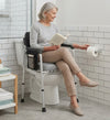 An older woman sits on a toilet seat risers with handles in a bright bathroom, reading a book while reaching for a toilet paper roll mounted on the wall.