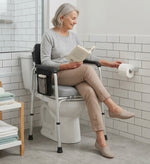 An older woman sits on a toilet seat risers with handles in a bright bathroom, reading a book while reaching for a toilet paper roll mounted on the wall.