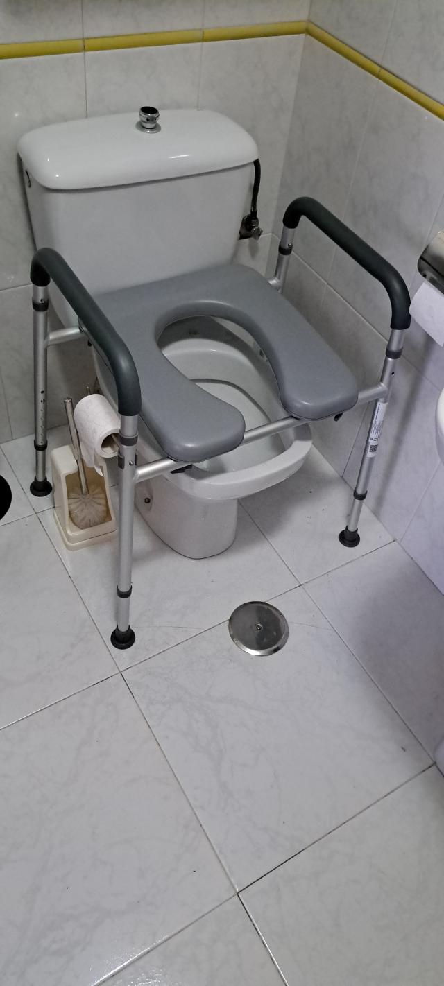 A gray and silver metal raised toilet lift seat with armrests, shown installed around a white toilet in a tiled bathroom.