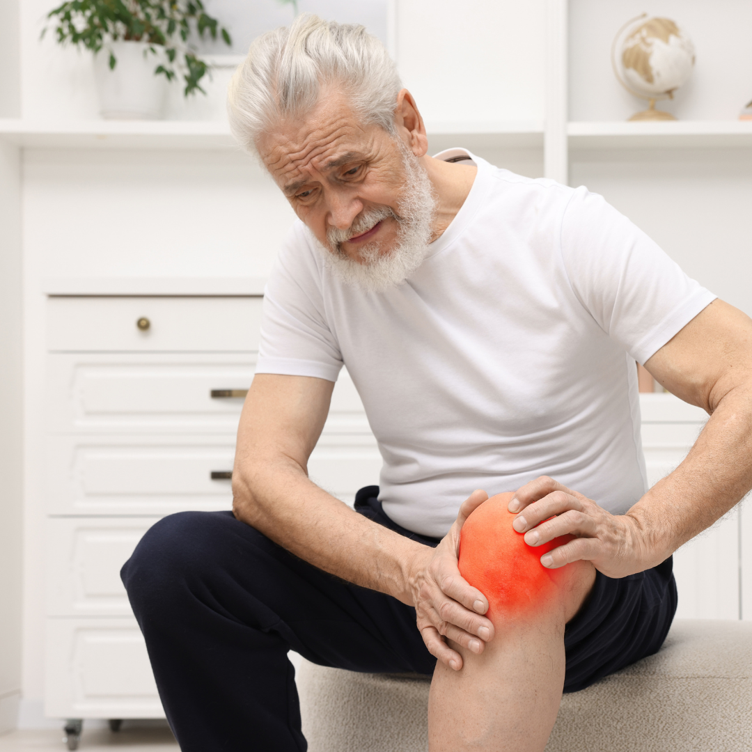 An older man with gray hair and a beard sits on a chair, holding his knee in pain. A glowing red heat effect is overlaid on his knee to indicate discomfort.
