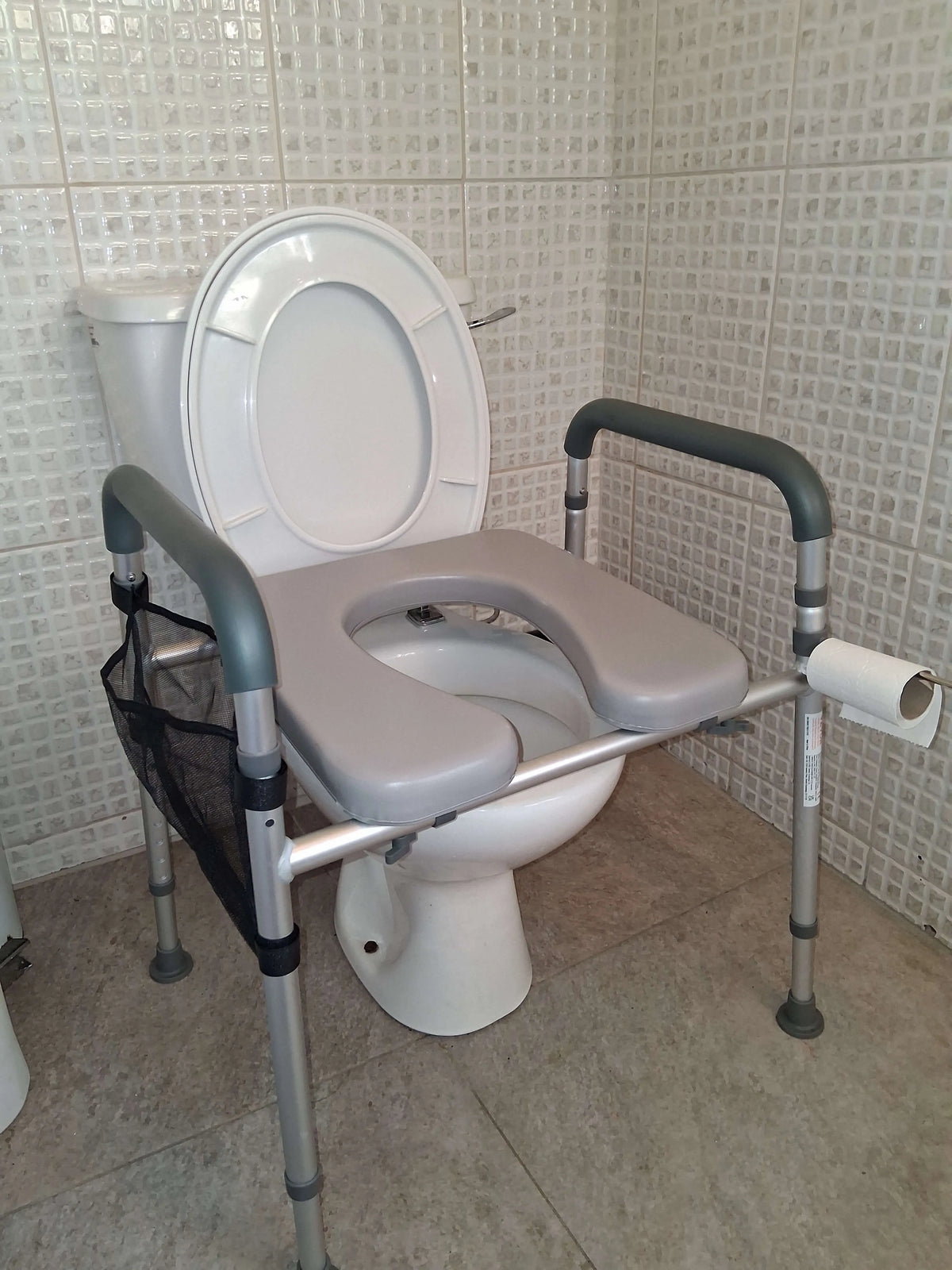 A gray and silver metal raised toilet seat and side rails with a black mesh storage bag, installed on a white toilet in a tiled bathroom.
