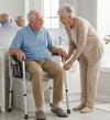 An elderly man sits on a raised toilet trainer chair with armrests, while an elderly woman in beige clothing assists him using a walker in a bright, modern bathroom.