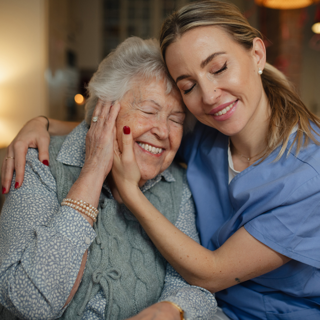 A smiling elderly woman in a patterned blue-gray sweater is embraced and comforted by a younger woman in blue scrubs, with warm indoor lighting and a softly blurred background.