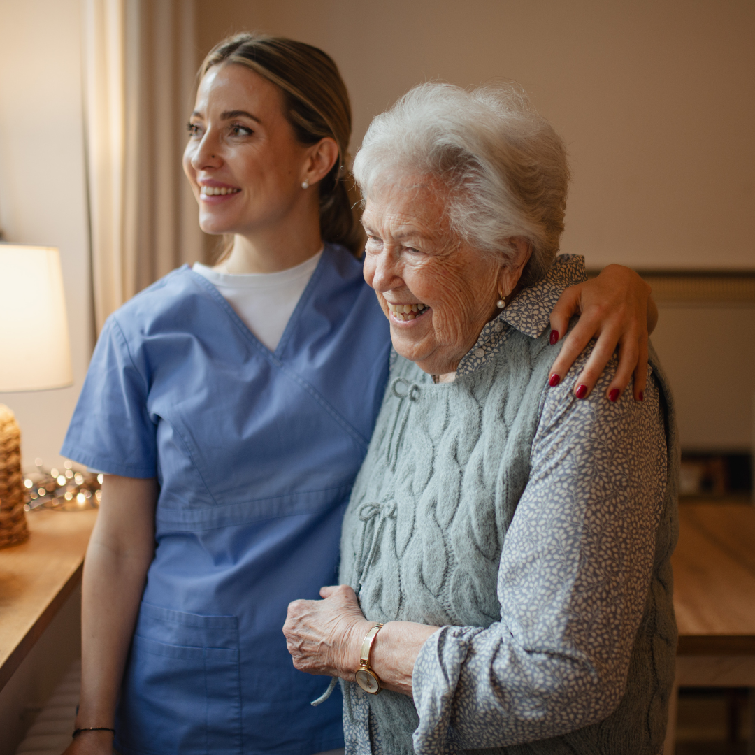 A smiling caregiver in blue scrubs stands beside an elderly woman wearing a gray cable-knit sweater, with the caregiver's arm around the older woman in a warmly lit indoor setting.