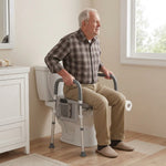 An older man sits on a white toilet with a gray toilet riser with armrests in a bright bathroom. The toilet riser seat has adjustable legs and side supports, and the man is wearing a brown plaid shirt and khaki pants.