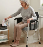 An older woman sits on a toilet seat riser with a portable sit-to-stand height-adjustable feature. The white toilet seat riser has black armrests with a mesh side pocket holding a magazine, and the word 'MONA' is visible on the leg.